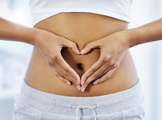 There is a woman wearing grey sweat pants and a cropped white top and holding her hands in a heart shape in front of her stomach.