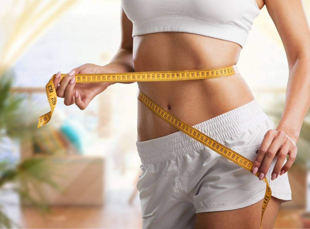 There is a woman in a white sports bra and white shorts. She is holding a yellow measuring tape around her waist.