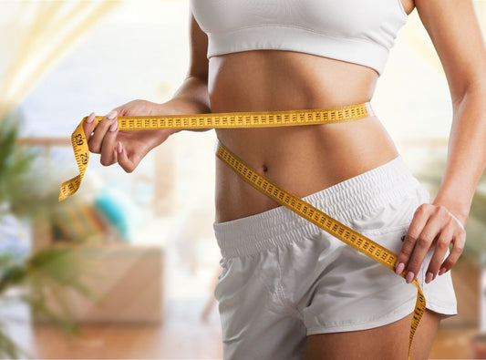 There is a woman in a white sports bra and white shorts. She is holding a yellow measuring tape around her waist.