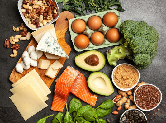 There are healthy foods like eggs, broccoli, and salmon on a black table top. There's also a wooden cutting board.