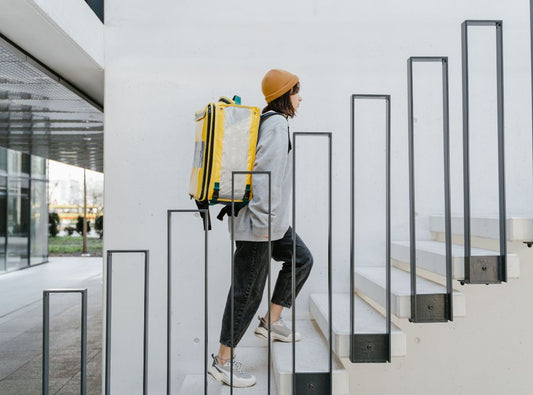 There is a woman in a grey sweater and black pants and an orange beanie with a yellow backpack climbing up stairs outdoors. Her sneakers are grey.