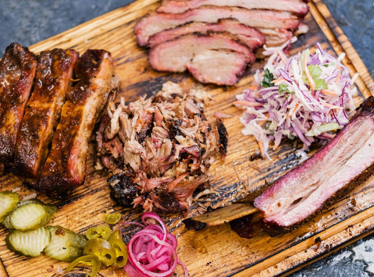 There are a selection of smoked meats and coleslaw on a wood cutting board. There are also pickles, peppers, and pickled onions.