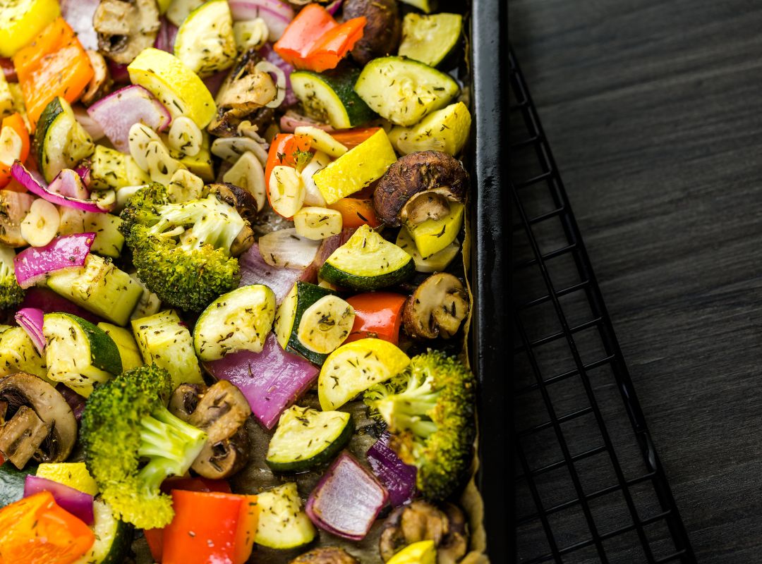 There is a tray of roasted vegetables. There are mushrooms, broccoli, onions, peppers, squash and other vegetables. The tray is on a gray table and has a black grid thing underneath it.