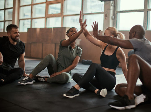 There is a group of people laughing on the floor in a gym. The left guy is wearing black, the left middle woman is wearing green clothes, the right woman is wearing black workout clothes, and the right man is wearing grey clothes.