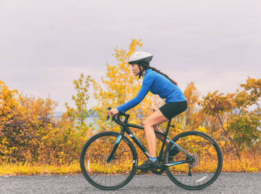 There is a woman with a blue shirt and black biking shorts on a black and blue bike. She is on a paved path with yellow leaves and a grey sky behind her.