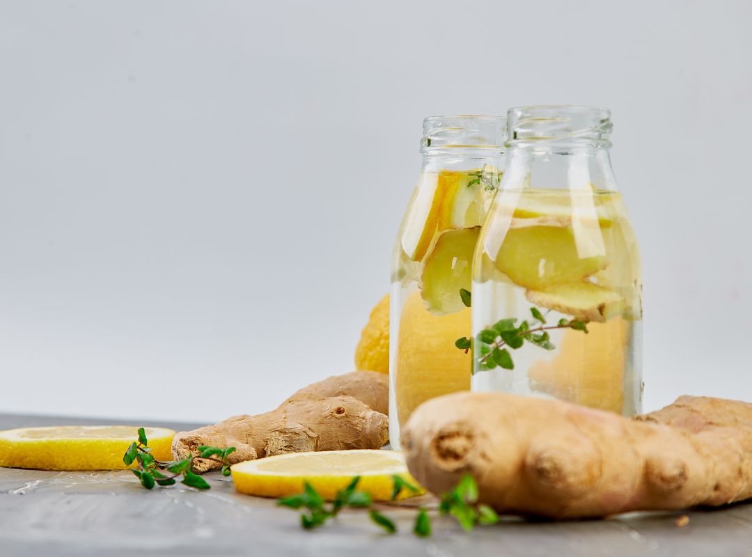 There are two glasses of lemon ginger water. Around the two glasses are pieces of lemon and ginger. There is also a green herb and all these things are on a grey table and there's a light grey background