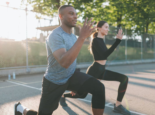 There is a man in blue and black sports clothes and a woman in black workout clothes doing a lunge outside.